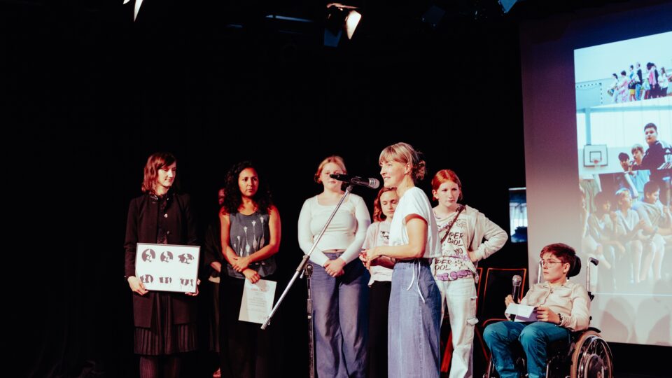 Eine Frau Am Mikrofon verleiht den dahinterstehenden Menschen einen der DIALOGE-Preise auf einer Bühne
