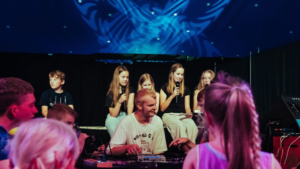 4 Mädchen im Hintergrund stehen auf einer Bühne und sprechen in Mikrophone, davor sitzt ein Junge an Synthesizern und schaut lächelnd Richtung Publikum