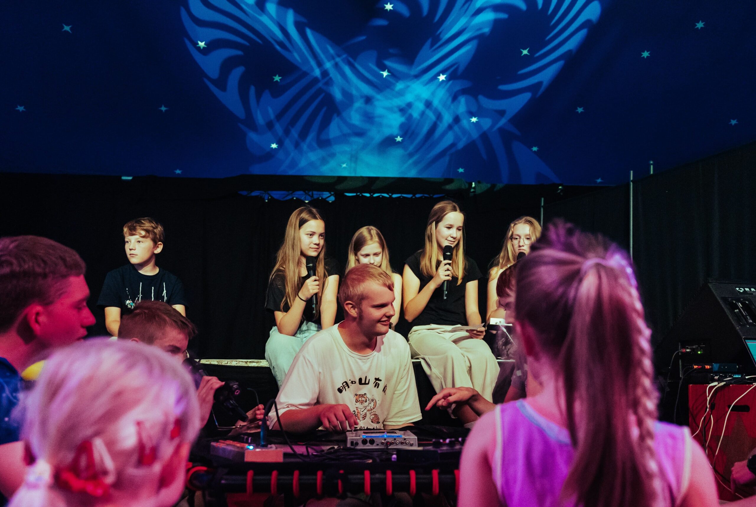 4 Mädchen im Hintergrund stehen auf einer Bühne und sprechen in Mikrophone, davor sitzt ein Junge an Synthesizern und schaut lächelnd Richtung Publikum